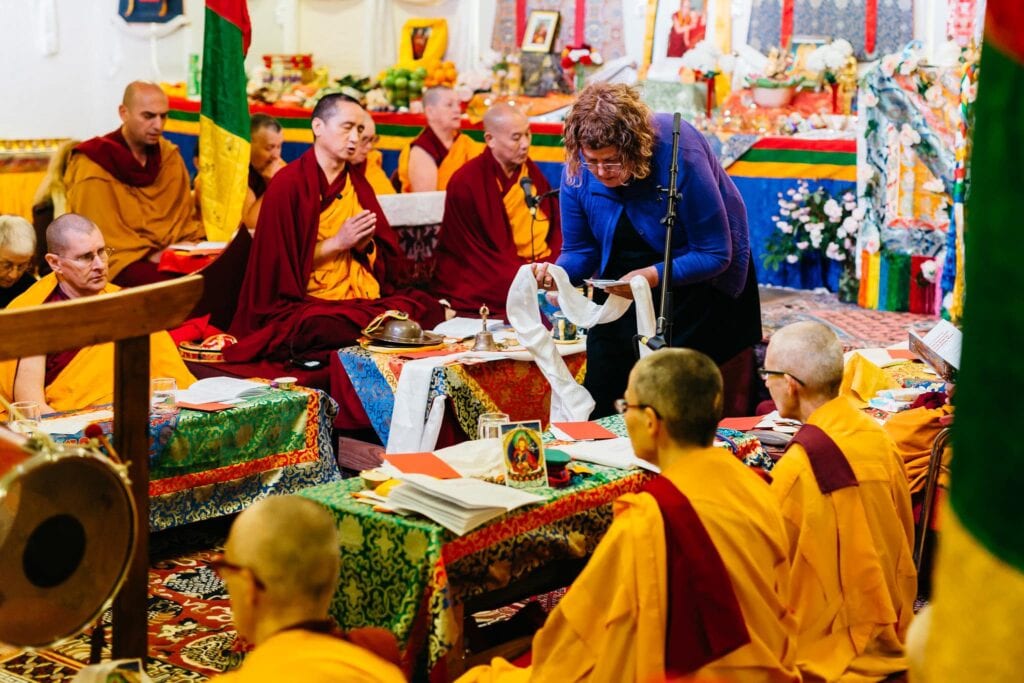 Monks and nuns seated for a puja while an offering scarf is presented